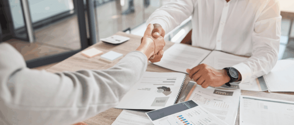 A imagem mostra duas pessoas se cumprimentando no momento de fechar um acordo, representando empresas de comércio B2B já adaptadas para os impactos e oportunidades da Reforma Tributária. Elas estão sentadas frente a frente, cada uma de um lado da mesa. Sobre a mesa tem vários papéis com textos, gráficos e tabelas.