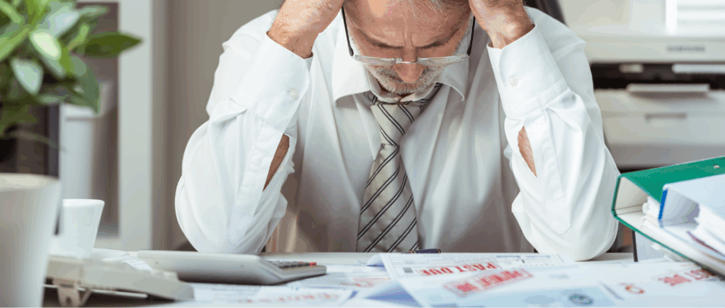 A imagem mostra um homem sentado em frente a uma mesa, com as mãos na cabeça em sinal de preocupação, representando um empresário que não se adequou à Reforma Tributária e seu negócio está prestes a quebrar. Sobre a mesa há várias contas com carimbos vermelhos que passaram do prazo de pagamento.