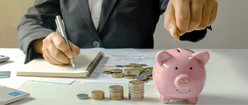 A imagem mostra um empresário analisando seus negócios. Ele está escrevendo em um caderno. Sobre a mesa ainda tem alguns papéis com gráficos, várias moedas e um porquinho rosa, no qual ele coloca uma moeda, representando o empresário que está prosperando na Reforma Tributária com a ajuda de seu contador.