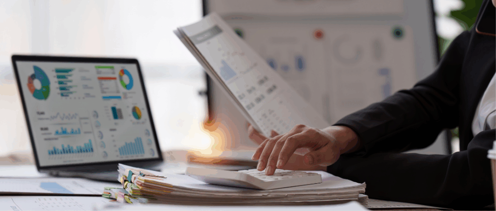 A imagem mostra o perfil de uma pessoa ao lado direito, em frente a uma mesa, representando a figura de um empresário analisando a Reforma Tributária em seu e-commerce. Na mesa tem um notebook com vários gráficos na tela, diversas pilhas de papéis e uma calculadora sobre eles. A pessoa está segurando alguns papéis com números e gráficos e fazendo contas na calculadora. Ao fundo, desfocado, tem um quadro branco com mais gráficos, representando números importantes do negócio de e-commerce.