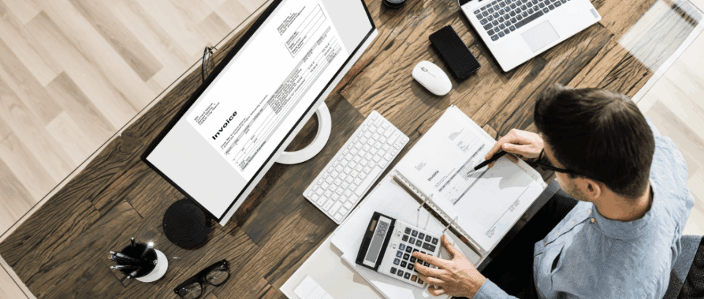A imagem mostra um empresário analisando documentos e fazendo cálculos com uma caneta e uma calculadora, representando uma empresa que está se preparando para a Reforma Tributária. Ela está sentado em frente a uma mesa, que tem um fichário com vários documentos, uma calculadora, um notebook, um celular, um mouse, um monitor, um teclado, um porta canetas e óculos.
