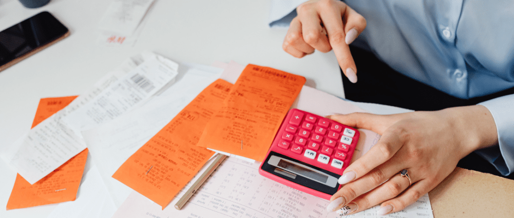 A imagem mostra uma pessoa fazendo contas em uma calculadora e analisando diversos papéis, representando a transformação que os registros empresariais terão no Brasil com o MAT, ou Módulo de Administração Tributária.