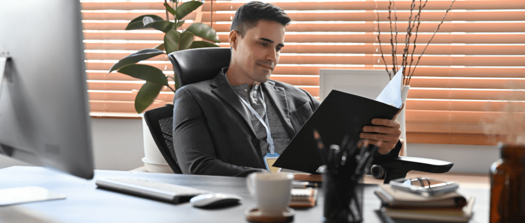 A imagem mostra um homem em seu escritório com uma mesa e, sobre ela, cadernos, lápis, uma caneca, um mouse, um teclado e um monitor. Ele está analisando os dados contábeis de seu negócio, representando um empresário que está verificando o que vai mudar para a sua empresa do Lucro Presumido com a Reforma Tributária.