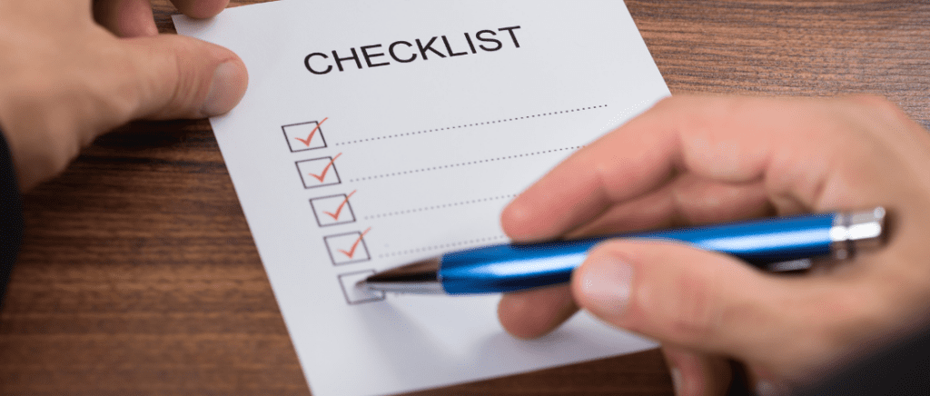 A imagem mostra as mãos de uma pessoa segurando uma caneta azul e um papel que está sobre uma mesa de madeira. Nele está escrita a palavra “checklist” e abaixo vários quadrados ticados seguidos por várias linhas pontilhadas, representando um empresário conferindo uma lista de pontos essenciais para se preparar para a Reforma Tributária.
