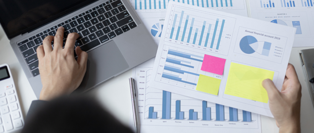 A imagem mostra a visão de cima da mesa de um empresário. A pessoa está analisando vários gráficos e mexendo no computador, representando o dono de um negócio que quer se adequar de modo correto.A imagem mostra a visão de cima da mesa de um empresário. A pessoa está analisando vários gráficos e mexendo no computador, representando o dono de um negócio que quer se adequar de modo correto.