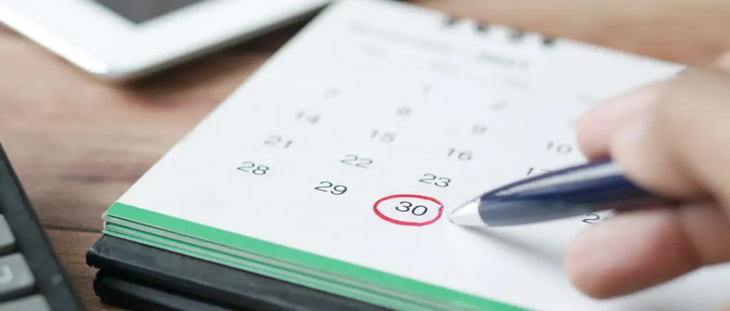 A imagem mostra uma calculadora, um tablet e um calendário de papel em cima de uma mesa de escritório. Além disso, tem uma pessoa segurando uma caneta, que circulou a data do dia 30, representando o empresário que está anotando as obrigações da agenda tributária de agosto de 2026.
