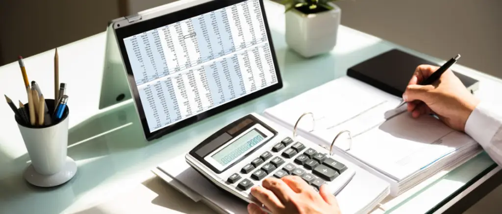 A imagem mostra o dono de uma empresa em sua mesa de trabalho. Sobre ela, tem um tablet com várias tabelas, um porta-canetas, um vaso pequeno com uma planta, um fichário aberto com uma calculadora. O empresário está fazendo cálculos e anotando na folha ao lado, representando as contas da alíquota-teste de 1% sobre o CBS e IBS em 2026.