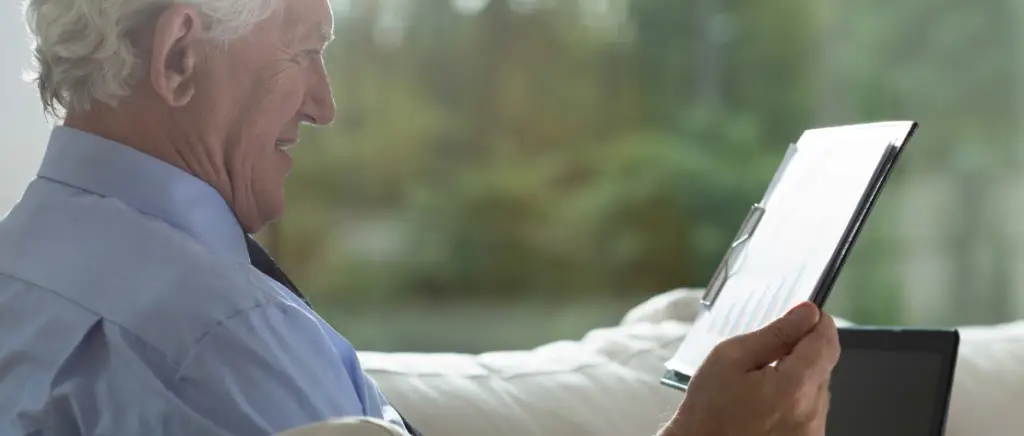 A imagem mostra um dono de um escritório contábil planejando a sucessão do seu negócio. Ele está sentado em um sofá com um notebook em seu colo e segurando uma prancheta com vários dados sobre o seu escritório de contabilidade.